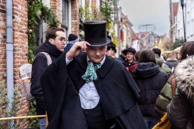 Deventer, Overijssel, Hollanda - 14.12.2025: Charles Dickens Festivali 'nde. Viktorya dönemi kostümlü adam, şenlikli caddede çelenklerle, süslerle ve seyircilerle silindir şapka takıyor..