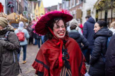 Deventer, Overijssel, Hollanda - 14.12.2025: Charles Dickens Festivali 'nde. Kırmızı pelerinli ve süslü şapkalı gülen şahıs, şenlikli sokak etkinliğinde kış kalabalığının ortasında duruyor..