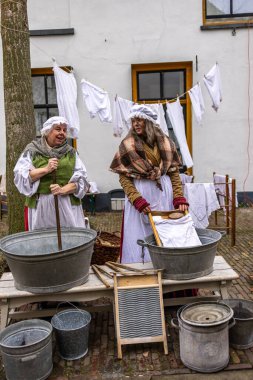 Deventer, Overijssel, Hollanda - 14.12.2025: Charles Dickens Festivali 'nde. Kapüşonlu ve önlüklü iki kadın geleneksel çamaşırlarını çamaşır küvetleri ve çamaşır ipleriyle gösteriyorlar..
