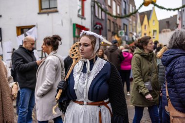 Deventer, Overijssel, Hollanda - 14.12.2025: Charles Dickens Festivali 'nde. Şapkalı ve önlüklü kadın, şenlikli sokak etkinliğinde çelenklerle ve kalabalıkla örülmüş personeli tutuyor..
