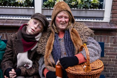 Deventer, Overijssel, Hollanda - 14.12.2025: Charles Dickens Festivali 'nde. Katmanlı eski giysiler içinde iki kişi tiyatro sahnesinde sepet ve şişeyle bir bankta yan yana oturur..