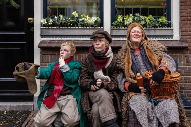 Deventer, Overijssel, Hollanda - 14.12.2025: Charles Dickens Festivali 'nde. Eski püskü giysiler içinde üç kişi, tiyatro sahnesinde sahne donanımlarıyla bankta poz veriyor..