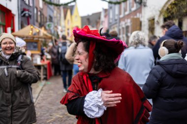 Deventer, Overijssel, Hollanda - 14.12.2025: Charles Dickens Festivali 'nde. Kırmızı pelerinli ve süslü şapkalı gülen şahıs, şenlikli sokak etkinliğinde kış kalabalığının ortasında duruyor..