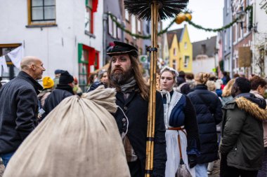 Deventer, Overijssel, Hollanda - 14.12.2025: Charles Dickens Festivali 'nde. Baca fırçalı sakallı adam ve kapüşonlu hizmetçi süslü binalarla dolu bir sokak etkinliğinde poz veriyorlar..