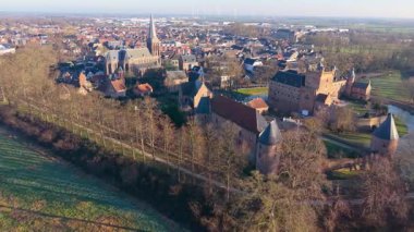 Yüksek bir kilise kulesi, güçlendirilmiş kale, kırmızı çatı evleri, yeşil tarlalar ve uzak rüzgar türbinlerinin canlı bir miras ve modern sürdürülebilirlik karışımı oluşturduğu bir Avrupa kasabasının havadan görünüşü.