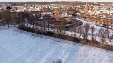 Karla kaplı bir kasaba, içinde kuleler ve hendek kış manzarası olan kırmızı tuğladan bir kale, kilise kulesi, çatılar ve ağaçlar sakin bir hava manzarası yaratır..