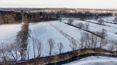 Karlı kırsal alan, dağınık yapraksız ağaçlar ve uzak kasaba çan kulesinin yumuşak ışığı ve bulutlu gökyüzü ile huzurlu bir kış ortamı yaratıyor..