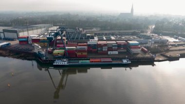 Yığınlanmış kargo konteynırlarıyla dolu büyük bir liman ve nehir boyunca yoğun bir sanayi sahnesi oluşturan vinçlerle, kamyonlarla ve depolarla dolu bir liman..