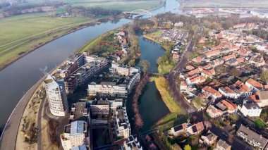 Çağdaş binalar, buhar yayan yüksek eğimli çatı kulesi, kenetlenmiş tekneler ve çevreleyen yeşil alanlar temiz bir yarı şehir manzarası oluşturan modern bir nehir kenarı bölgesi..