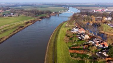 Kargo gemisi, kemerli köprü ve karışık kullanım bankaları olan nehrin havadan görüntüsü. Yerleşim evleri, bahçeler ve yeşil tarlalar, kırsal yaşam ve su aracılığı ile taşınan sakin bir manzara çerçevesi oluşturuyor..