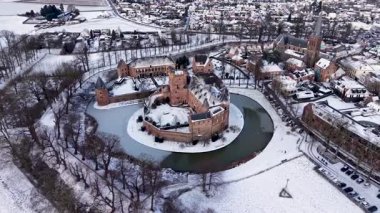 Kırmızı tuğladan kale ve donmuş ortaçağ kuleleriyle kaplı kar manzarası modern evlerle ve huzurlu kış manzarasındaki kilise kulesiyle tezat oluşturuyor..