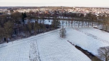 Karlı kırsal alan, dağınık yapraksız ağaçlar ve uzak kasaba çan kulesinin yumuşak ışığı ve bulutlu gökyüzü ile huzurlu bir kış ortamı yaratıyor..
