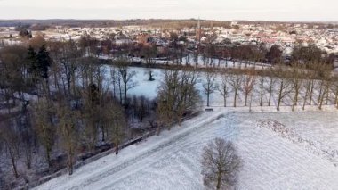 Karlı kırsal alan, dağınık yapraksız ağaçlar ve uzak kasaba çan kulesinin yumuşak ışığı ve bulutlu gökyüzü ile huzurlu bir kış ortamı yaratıyor..