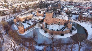 Kar, kuleleri, duvarları ve iç avluları olan kırmızı tuğladan kale kompleksi kış kasabasının çatıları, ağaçları ve çevreleyen binalar arasında durmaktadır..