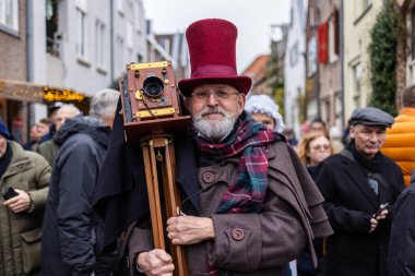 Deventer, Overijssel, Hollanda - 14.12.2025: Eski giyimli bir adam, canlı bir sokak etkinliği sırasında omuzunda antik bir ahşap kamera taşıyor.