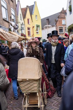 Deventer, Overijssel, Hollanda - 14.12.2025: Kürklü bir kadın, Viktorya dönemi kıyafetleri giymiş bir adamın yanında klasik bir bebek arabasını itiyor.