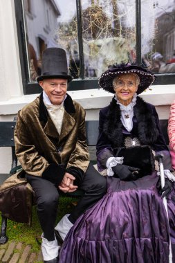Deventer, Overijssel, Hollanda - 14.12.2025: Viktorya döneminden kalma şık giyimli iki yaşlı insan, şenlikli ve süslü bir pencerenin önünde bir bankta oturuyor.