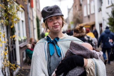 Deventer, Overijssel, Hollanda - 14.12.2025: Kaliteli giyimli bir kişi canlı ve kalabalık bir sokakta yürürken bir oyuncak bebek tutuyor.