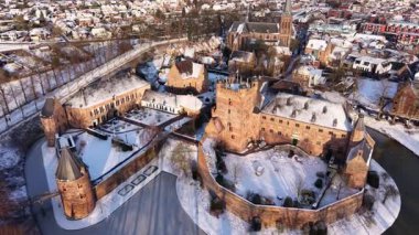 Bir hendek, yakınındaki binalar, yapraksız ağaçlar ve kış kırsalına uzanan küçük bir kasaba ile çevrili büyük tuğladan bir kaleyle kaplı bir kar manzarası..