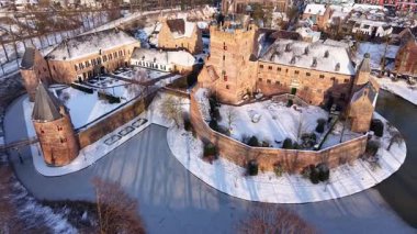 Bir hendek, yakınındaki binalar, yapraksız ağaçlar ve kış kırsalına uzanan küçük bir kasaba ile çevrili büyük tuğladan bir kaleyle kaplı bir kar manzarası..