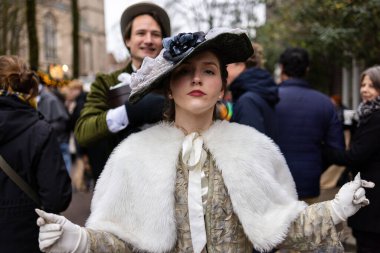 Deventer, Overijssel, Hollanda - 14.12.2025: Büyük bir şapka ve kürk atkısı olan zarif tarihi kıyafetli kadın, dönemin kostümlü bir erkeğin arkasında gülümsemesi gibi emin bir şekilde ayakta duruyor.