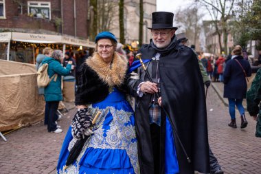 Süslü tarihi kostümler içinde iki kişi, biri güneş şemsiyeli mavi işlemeli bir elbise, diğeri de siyah pelerinli, festivalsel bir cadde pazarında yürüyor..