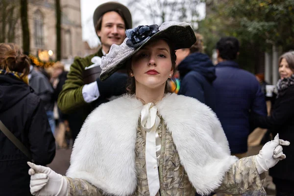Deventer, Overijssel, Hollanda - 14.12.2025: Büyük bir şapka ve kürk atkısı olan zarif tarihi kıyafetli kadın, dönemin kostümlü bir erkeğin arkasında gülümsemesi gibi emin bir şekilde ayakta duruyor.