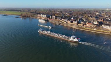 İki kargo gemisi, bir taş set, ikiz kule kilisesi olan tarihi binalar ve uzaklığa kadar uzanan çevresindeki tarım arazilerinin yer aldığı nehir kenarı manzarası..