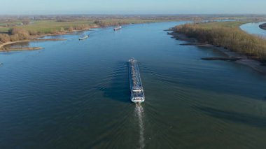 Etrafı düz yeşil bankalar ve kırsal alanda uzak rüzgar türbinleriyle çevrili birden fazla kargo gemisinin hareket ettiği geniş bir nehir..