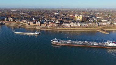 İki kargo gemisi, bir taş set, ikiz kule kilisesi olan tarihi binalar ve uzaklığa kadar uzanan çevresindeki tarım arazilerinin yer aldığı nehir kenarı manzarası..