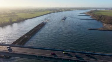 Bir kargo mavnası ve bir yolcu gemisi aşağıda yeşil tarlalar, ağaçlar ve uzak rüzgar türbinleriyle çevriliyken, yoğun bir köprünün üzerinden geçen geniş bir nehrin havadan görüntüsü..