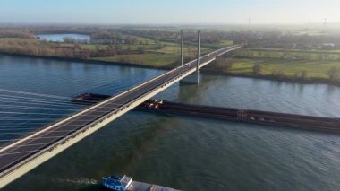 Aşağıdan geçen iki büyük gemi, yeşil tarlalar, orman arazileri ve sakin bir kırsal alanda uzak rüzgar türbinleri ile çevrili iken, bir kablo görüntüsü geniş bir nehir boyunca köprü olarak kaldı..