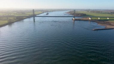 Geniş bir nehrin havadan görünüşü modern bir köprü ile kesişiyor, sakin su boyunca hareket eden bir mavna, yeşil tarlalar, seyrek ağaçlar ve kırsal alanda uzak rüzgar türbinleri ile çevrili..