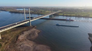 Aşağıdan geçen iki büyük gemi, yeşil tarlalar, orman arazileri ve sakin bir kırsal alanda uzak rüzgar türbinleri ile çevrili iken, bir kablo görüntüsü geniş bir nehir boyunca köprü olarak kaldı..