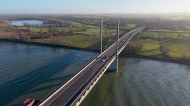 Aşağıdan geçen bir mavna, yeşil tarlalar, dağınık ağaçlar, küçük bir kasaba ve ufuktaki uzak rüzgar türbinleriyle çevrili, geniş bir nehri kaplayan bir köprü olarak kaldı..