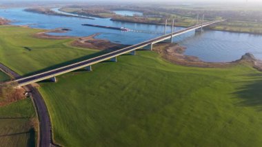 Kablo görüntüsü geniş bir nehir üzerinde köprü vazifesi gördü, aşağıda iki kargo gemisi, yeşil tarlalar, ağaçlar ve uzak rüzgar türbinleri sakin bir kırsal zemin oluşturuyordu..