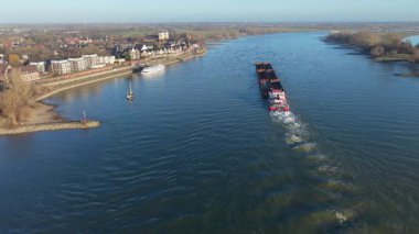Akıntıya karşı ilerleyen büyük bir yük mavnası olan bir nehrin havadan görünüşü. Evleri olan küçük bir kasabayı, rıhtımlı bir yolcu teknesini, bir yelkenliyi ve uzak rüzgar türbinleriyle çevrili tarlaları görüyoruz..
