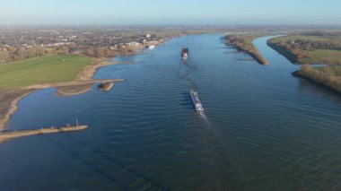 Durgun su boyunca hareket eden iki kargo gemisinin bulunduğu geniş bir nehrin hava manzarası. Çimenli tarlalar, küçük yerleşimler, ağaç yamaları ve temiz bir gökyüzünün altındaki kumsallarla çevrili..