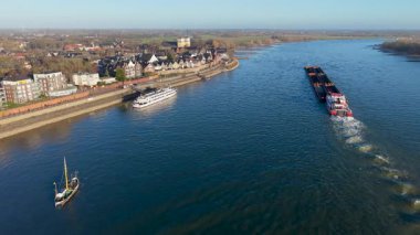 Akıntıya karşı ilerleyen büyük bir yük mavnası olan bir nehrin havadan görünüşü. Evleri olan küçük bir kasabayı, rıhtımlı bir yolcu teknesini, bir yelkenliyi ve uzak rüzgar türbinleriyle çevrili tarlaları görüyoruz..