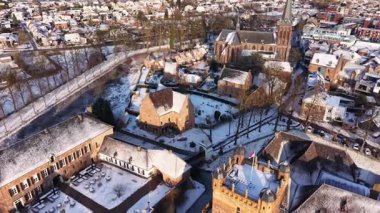 Merkezi bir kulesi, kırmızı panjurları ve etrafındaki tuğla binaları olan karla kaplı bir şatonun hava manzarası bir kilise ve sıcak güneş ışığıyla parlayan tarihi bir kasabanın yanında geçiyor..