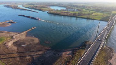 Halatın altındaki dolambaçlı bir nehir boyunca bir mavnayı iten römorkör görüntüsü çimenli tarlarla çevrili kalmış nehir kıyıları ve açık havada dağılmış ağaçlar..