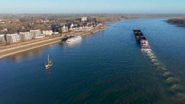 Büyük bir kargo gemisinin akıntı yönünde ilerlediği bir nehrin havadan görüntüsü, kıyıya yanaşmış küçük tekneler, kıyı şeridinde bir kasaba, ve uzak mesafede rüzgar türbinleri olan açık alanlar..