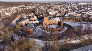 Karla kaplı kırmızı tuğlalı kale, tarihi bir kasaba, kilise kulesi, tarlalar ve ormanlar tarafından çevrelenmiş bir hendek ve kuleler..