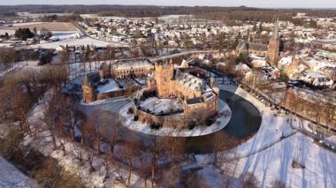 Karla kaplı kırmızı tuğlalı kale, tarihi bir kasaba, kilise kulesi, tarlalar ve ormanlar tarafından çevrelenmiş bir hendek ve kuleler..