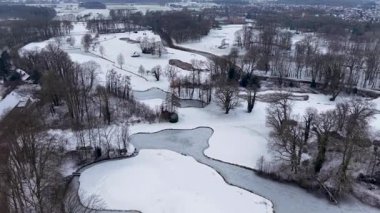 Yapraksız ağaçlar, dolambaçlı yollar ve yakınlardaki bir kasaba tarafından çevrili donmuş göletler ve karla kaplı hava manzarası sakin ve geniş bir kış manzarası yaratıyor..