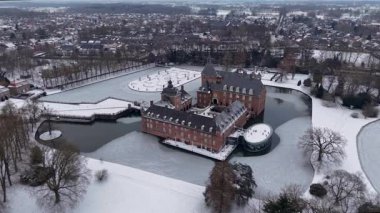 Kızıl tuğladan bir şatonun havadan görünüşü, kuleleri ve hendeği olan, karla kaplı resmi bir bahçenin yanında, ağaçlarla çevrili ve yakınlardaki bir kasaba, dingin, beyaz bir manzara..