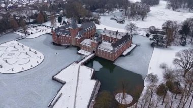 Yapraksız ağaçlar ve açık karlı tarlalarla çevrili kar kaplı resmi bir bahçenin yanında, kuleleri ve kısmen donmuş hendeği olan kırmızı tuğladan bir kalenin havadan görünüşü..