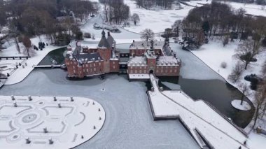 Yapraksız ağaçlar ve açık karlı tarlalarla çevrili kar kaplı resmi bir bahçenin yanında, kuleleri ve kısmen donmuş hendeği olan kırmızı tuğladan bir kalenin havadan görünüşü..