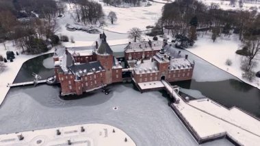 Yapraksız ağaçlar ve açık karlı tarlalarla çevrili kar kaplı resmi bir bahçenin yanında, kuleleri ve kısmen donmuş hendeği olan kırmızı tuğladan bir kalenin havadan görünüşü..