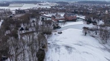 Karlı tarlalar, donmuş su yolları, yapraksız ağaçlar ve arazi boyunca uzanan uzak bir kasaba ile çevrili kuleleri ve hendeği olan büyük kırmızı tuğladan bir kalenin havadan görünüşü..